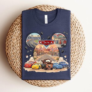 Puede incluir: Una camiseta azul marino con un estampado gr&aacute;fico de los personajes de Disney Pixar Cars, incluyendo a Rayo McQueen y Mate. El dise&ntilde;o incluye un marco con forma de orejas de Mickey que muestra escenas de una pista y Radiator Springs.