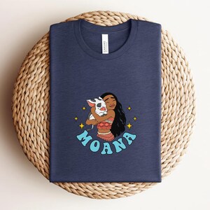 Puede incluir: Una camiseta azul marino de cuello redondo con una ilustraci&oacute;n gr&aacute;fica de un personaje de pelo largo y oscuro abrazando a un peque&ntilde;o cerdo manchado. El dise&ntilde;o incluye estrellas amarillas de cuatro puntas y el texto MOANA en letras may&uacute;sculas azul claro.