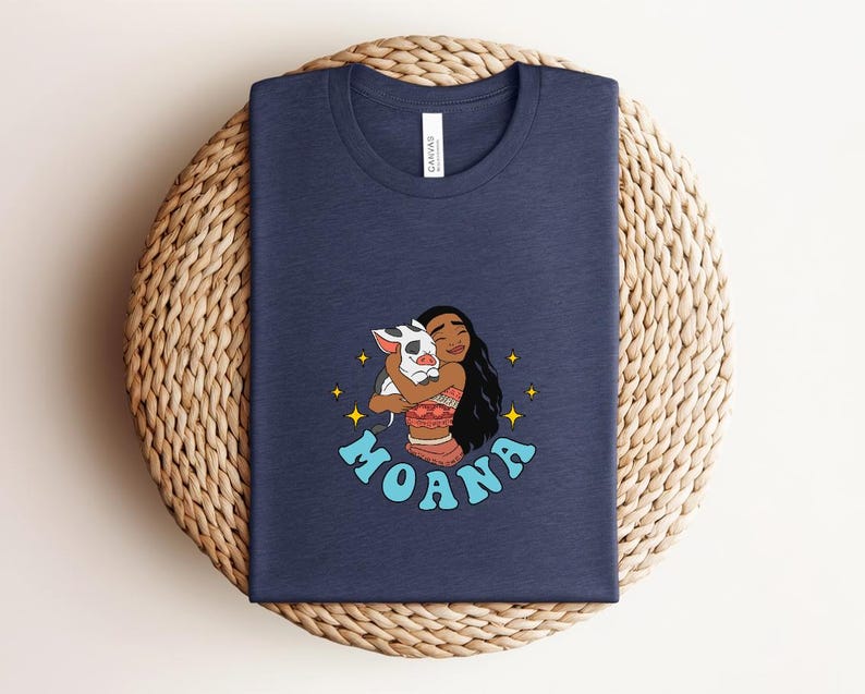 Puede incluir: Una camiseta azul marino de cuello redondo con una ilustraci&oacute;n gr&aacute;fica de un personaje de pelo largo y oscuro abrazando a un peque&ntilde;o cerdo manchado. El dise&ntilde;o incluye estrellas amarillas de cuatro puntas y el texto MOANA en letras may&uacute;sculas azul claro.