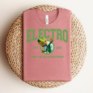 May include: A dusty rose t-shirt with the word "ELECTRO" in green block letters. Below the word is a cartoon image of a character in green and yellow. The text "EST 2021" and "SPIDEY AND HIS AMAZING FRIENDS" are also printed on the shirt.