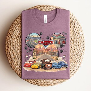 Puede incluir: Una camiseta malva de cuello redondo con un estampado gr&aacute;fico del logotipo de Disney Pixar Cars sobre tres escenas circulares de un circuito, una gasolinera y un pueblo del desierto. Debajo, cinco coches animados aparecen en la carretera.