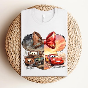May include: A white t-shirt featuring a graphic of a bow shape containing Tow Mater on the left and Lightning McQueen on the right. The left side is brown and rustic, while the right side is bright red and orange. The names Tow Mater and Lightning McQueen are printed below the characters.