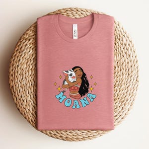 Puede incluir: Una camiseta de cuello redondo en color rosa empolvado con un gr&aacute;fico del personaje Moana abrazando a un cerdito. El dise&ntilde;o incluye estrellas amarillas y el nombre MOANA en letras azul claro. La camiseta tiene una etiqueta blanca en el cuello.
