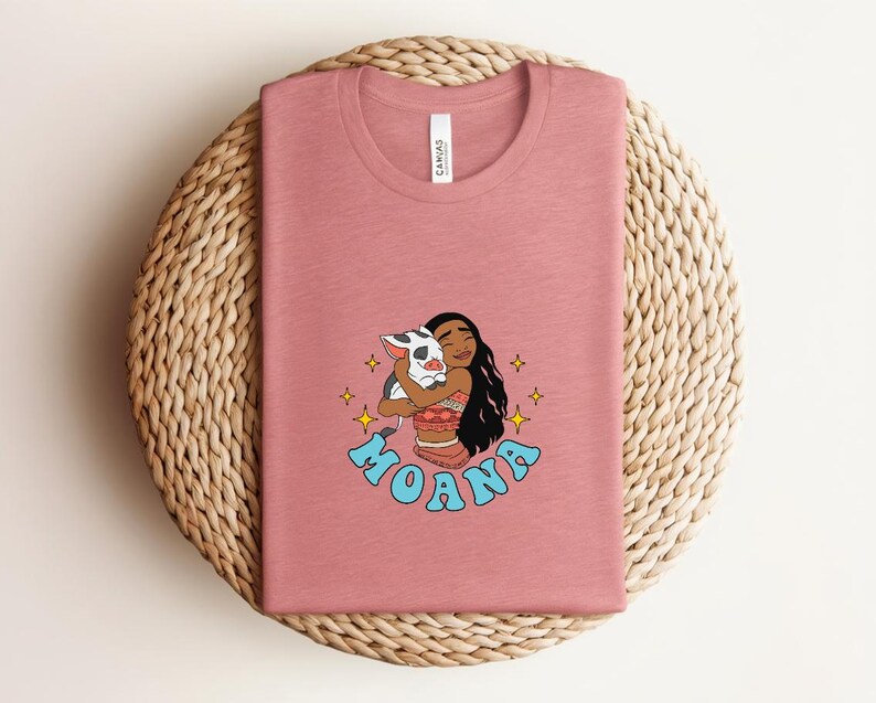 Puede incluir: Una camiseta de cuello redondo en color rosa empolvado con un gr&aacute;fico del personaje Moana abrazando a un cerdito. El dise&ntilde;o incluye estrellas amarillas y el nombre MOANA en letras azul claro. La camiseta tiene una etiqueta blanca en el cuello.
