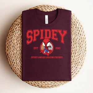 May include: A maroon t-shirt featuring the word "SPIDEY" in red block letters above a cartoon image of Spider-Man. The text "EST 2021" and "SPIDEY AND HIS AMAZING FRIENDS" are also printed on the shirt. The shirt is displayed on a woven, circular surface.
