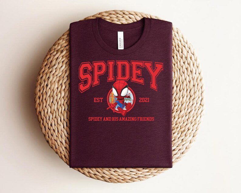 May include: A maroon t-shirt featuring the word "SPIDEY" in red block letters above a cartoon image of Spider-Man. The text "EST 2021" and "SPIDEY AND HIS AMAZING FRIENDS" are also printed on the shirt. The shirt is displayed on a woven, circular surface.