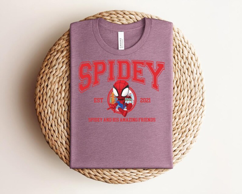 May include: A mauve t-shirt featuring a red graphic of Spider-Man with the text "SPIDEY" above and "EST 2021" and "SPIDEY AND HIS AMAZING FRIENDS" below. The shirt is displayed on a woven, circular mat.