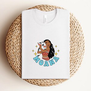 Puede incluir: Una camiseta blanca con una ilustraci&oacute;n gr&aacute;fica de un personaje de pelo largo y oscuro abrazando a un peque&ntilde;o lech&oacute;n. El dise&ntilde;o incluye estrellas amarillas de cuatro puntas y la palabra MOANA en letras de burbuja azul claro debajo.