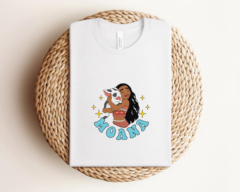 Puede incluir: Una camiseta blanca con una ilustraci&oacute;n gr&aacute;fica de un personaje de pelo largo y oscuro abrazando a un peque&ntilde;o lech&oacute;n. El dise&ntilde;o incluye estrellas amarillas de cuatro puntas y la palabra MOANA en letras de burbuja azul claro debajo.
