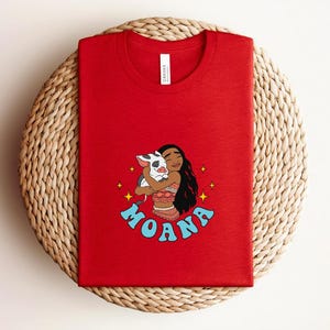 Puede incluir: Una camiseta roja de cuello redondo con un estampado gr&aacute;fico de un personaje de pelo largo y oscuro abrazando a un cerdito. El dise&ntilde;o incluye estrellas amarillas y el texto azul MOANA impreso en una fuente curva bajo la ilustraci&oacute;n.