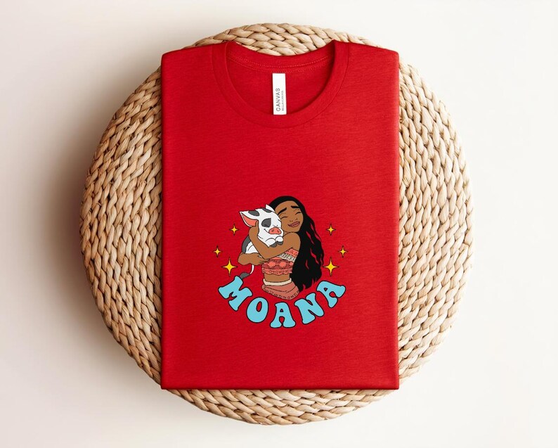 Puede incluir: Una camiseta roja de cuello redondo con un estampado gr&aacute;fico de un personaje de pelo largo y oscuro abrazando a un cerdito. El dise&ntilde;o incluye estrellas amarillas y el texto azul MOANA impreso en una fuente curva bajo la ilustraci&oacute;n.