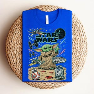 Puede incluir: Una camiseta azul brillante con un estampado gr&aacute;fico de Grogu de Star Wars, rodeado de varias naves espaciales, la Estrella de la Muerte, BB-8 y R2-D2 sobre una superficie con cr&aacute;teres. El texto STAR WARS aparece en letras negras grandes.