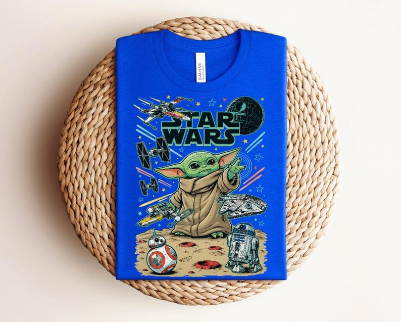 Puede incluir: Una camiseta azul brillante con un estampado gr&aacute;fico de Grogu de Star Wars, rodeado de varias naves espaciales, la Estrella de la Muerte, BB-8 y R2-D2 sobre una superficie con cr&aacute;teres. El texto STAR WARS aparece en letras negras grandes.