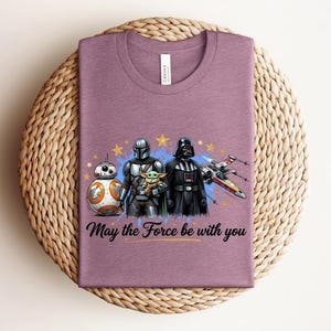 Pode incluir: Uma t-shirt malva de gola redonda com um gr&aacute;fico de BB-8, o Mandaloriano, Grogu e Darth Vader com um ca&ccedil;a X-wing. O design inclui estrelas amarelas, salpicos de tinta azul e o texto preto May the Force be with you.
