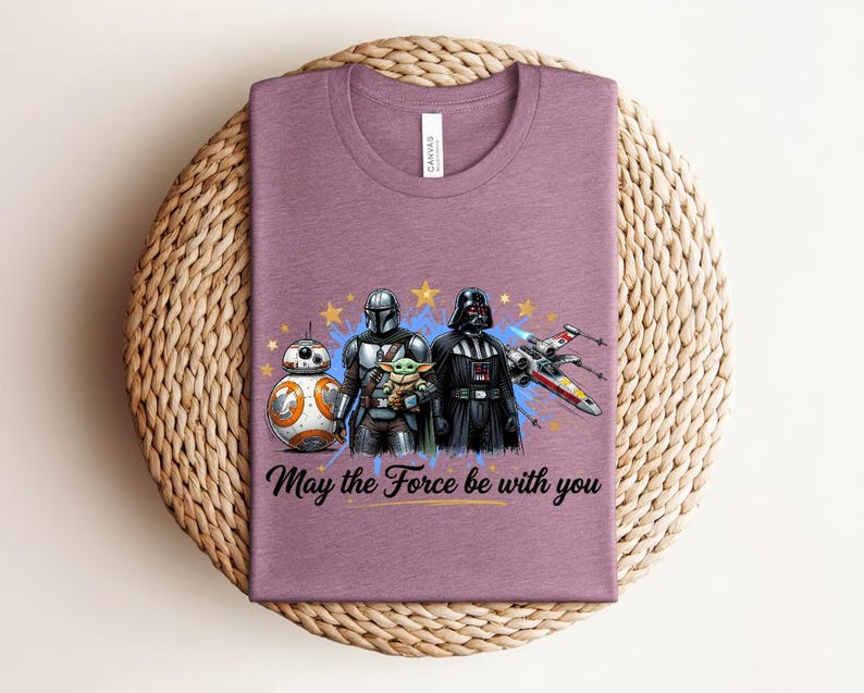 Pode incluir: Uma t-shirt malva de gola redonda com um gr&aacute;fico de BB-8, o Mandaloriano, Grogu e Darth Vader com um ca&ccedil;a X-wing. O design inclui estrelas amarelas, salpicos de tinta azul e o texto preto May the Force be with you.
