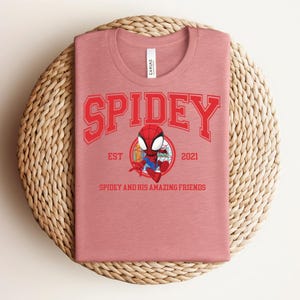May include: A dusty rose t-shirt featuring the word "SPIDEY" in red block letters above a cartoon Spider-Man graphic. Below the graphic are the words "EST 2021" and "SPIDEY AND HIS AMAZING FRIENDS" in red. The shirt is folded on a woven, circular placemat.
