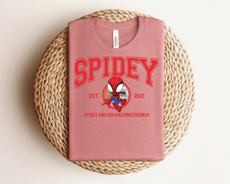 May include: A dusty rose t-shirt featuring the word "SPIDEY" in red block letters above a cartoon Spider-Man graphic. Below the graphic are the words "EST 2021" and "SPIDEY AND HIS AMAZING FRIENDS" in red. The shirt is folded on a woven, circular placemat.