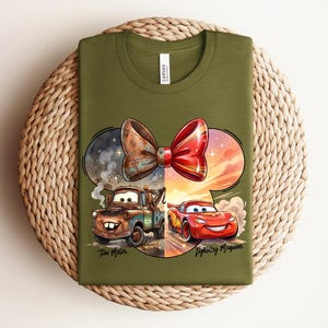 May include: An olive green t-shirt featuring a graphic of a red and brown bow shape containing illustrations of the characters Tow Mater and Lightning McQueen. The text "Tow Mater" and "Lightning McQueen" is printed below the characters.