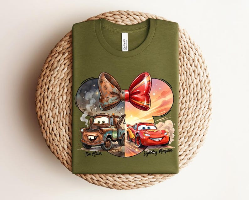 May include: An olive green t-shirt featuring a graphic of a red and brown bow shape containing illustrations of the characters Tow Mater and Lightning McQueen. The text "Tow Mater" and "Lightning McQueen" is printed below the characters.