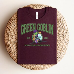 May include: A maroon t-shirt featuring the text "GREEN GOBLIN" in green, arched lettering. Below the text is a cartoon image of the Green Goblin character, with the text "EST 2021" and "SPIDEY AND HIS AMAZING FRIENDS". The shirt is displayed on a woven surface.