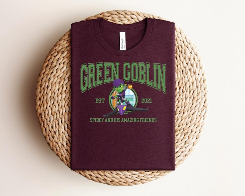 May include: A maroon t-shirt featuring the text "GREEN GOBLIN" in green, arched lettering. Below the text is a cartoon image of the Green Goblin character, with the text "EST 2021" and "SPIDEY AND HIS AMAZING FRIENDS". The shirt is displayed on a woven surface.
