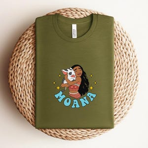 Puede incluir: Una camiseta verde oliva con una ilustraci&oacute;n gr&aacute;fica de un personaje de pelo largo y oscuro abrazando a un peque&ntilde;o cerdo blanco y negro. El dise&ntilde;o incluye estrellas amarillas de cuatro puntas y la palabra MOANA en letras azul claro.