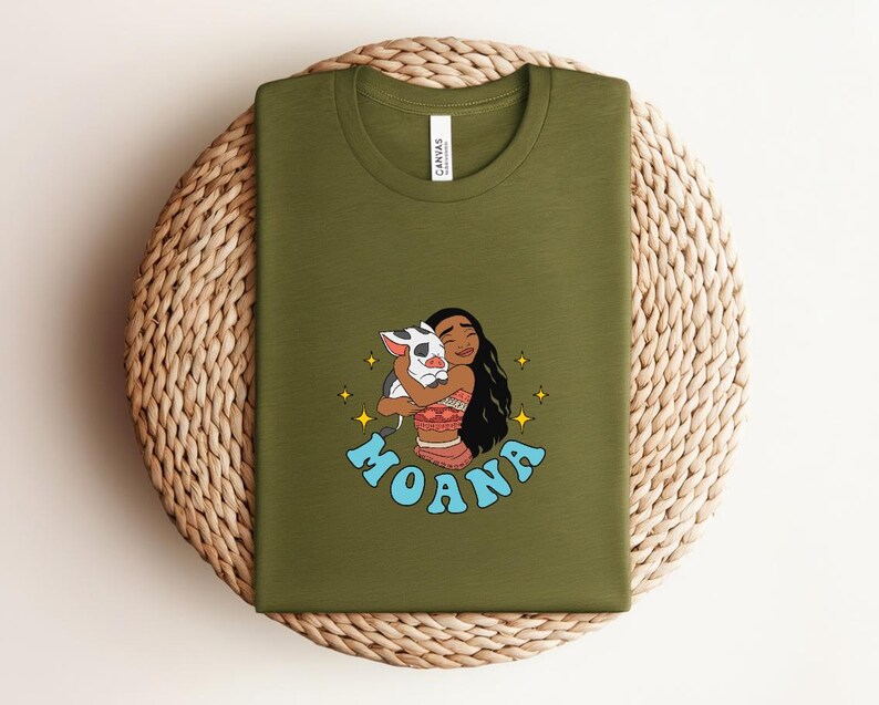 Puede incluir: Una camiseta verde oliva con una ilustraci&oacute;n gr&aacute;fica de un personaje de pelo largo y oscuro abrazando a un peque&ntilde;o cerdo blanco y negro. El dise&ntilde;o incluye estrellas amarillas de cuatro puntas y la palabra MOANA en letras azul claro.