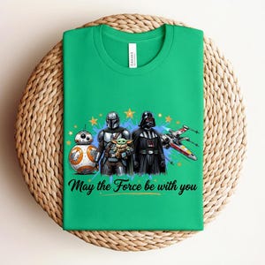 Pode incluir: Uma t-shirt verde brilhante com uma impress&atilde;o gr&aacute;fica de BB-8, O Mandaloriano, Grogu e Darth Vader ao lado de um ca&ccedil;a estelar X-wing. O design inclui estrelas amarelas, salpicos de tinta azul e o texto May the Force be with you.