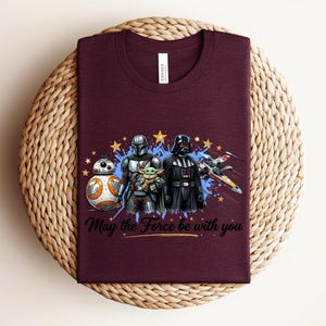 Pode incluir: Uma t-shirt de gola redonda cor de vinho com uma impress&atilde;o gr&aacute;fica de personagens de Star Wars, incluindo BB-8, O Mandaloriano, Grogu e Darth Vader, um ca&ccedil;a X-wing, estrelas amarelas, salpicos de tinta azul e o texto May the Force be with you.