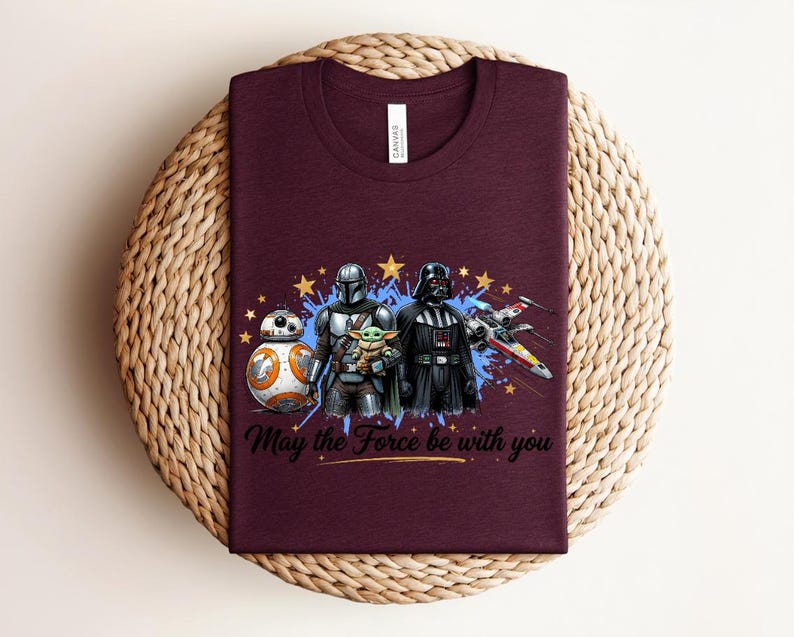 Pode incluir: Uma t-shirt de gola redonda cor de vinho com uma impress&atilde;o gr&aacute;fica de personagens de Star Wars, incluindo BB-8, O Mandaloriano, Grogu e Darth Vader, um ca&ccedil;a X-wing, estrelas amarelas, salpicos de tinta azul e o texto May the Force be with you.