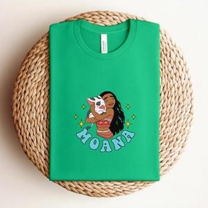 Puede incluir: Una camiseta verde brillante de manga corta con una ilustraci&oacute;n gr&aacute;fica de un personaje de pelo largo y oscuro abrazando a un peque&ntilde;o cerdo manchado. La palabra MOANA est&aacute; impresa en letras azules debajo del personaje, rodeada de estrellas amarillas.