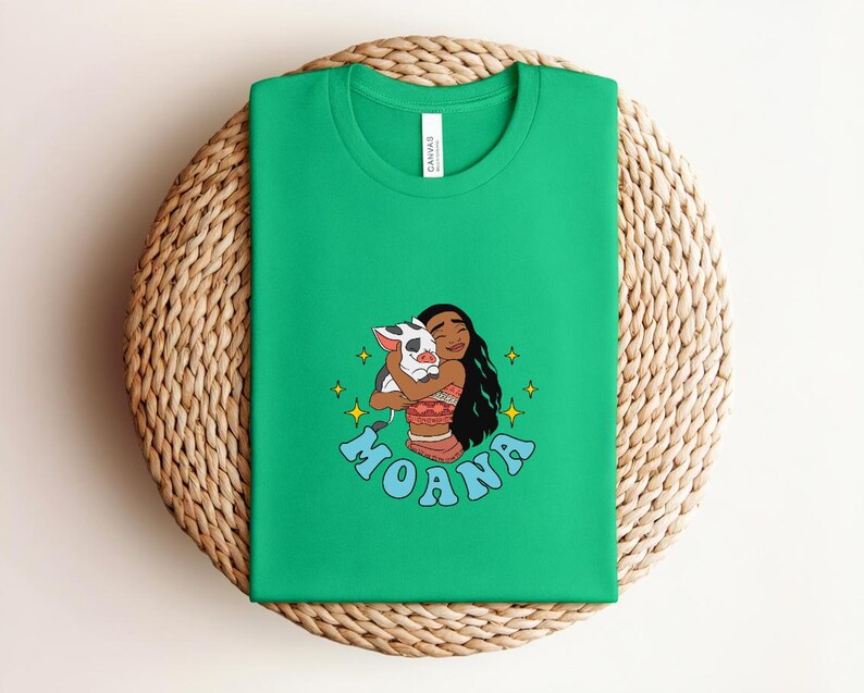 Puede incluir: Una camiseta verde brillante de manga corta con una ilustraci&oacute;n gr&aacute;fica de un personaje de pelo largo y oscuro abrazando a un peque&ntilde;o cerdo manchado. La palabra MOANA est&aacute; impresa en letras azules debajo del personaje, rodeada de estrellas amarillas.
