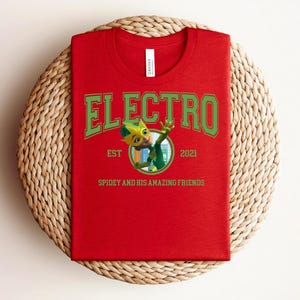 May include: A red t-shirt with the word "ELECTRO" in green block letters. Below the word is a cartoon character in a circle, with the text "EST 2021" and "SPIDEY AND HIS AMAZING FRIENDS". The shirt is on a woven mat.