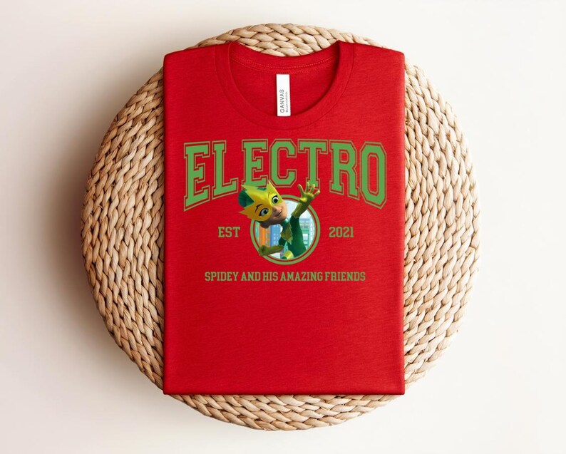 May include: A red t-shirt with the word "ELECTRO" in green block letters. Below the word is a cartoon character in a circle, with the text "EST 2021" and "SPIDEY AND HIS AMAZING FRIENDS". The shirt is on a woven mat.