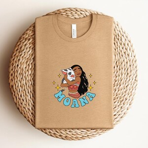 Puede incluir: Una camiseta de cuello redondo color canela con una ilustraci&oacute;n gr&aacute;fica de un personaje de pelo largo y oscuro abrazando a un peque&ntilde;o cerdo manchado. El dise&ntilde;o incluye estrellas amarillas de cuatro puntas y la palabra MOANA en letras azul claro.