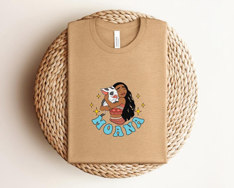 Puede incluir: Una camiseta de cuello redondo color canela con una ilustraci&oacute;n gr&aacute;fica de un personaje de pelo largo y oscuro abrazando a un peque&ntilde;o cerdo manchado. El dise&ntilde;o incluye estrellas amarillas de cuatro puntas y la palabra MOANA en letras azul claro.