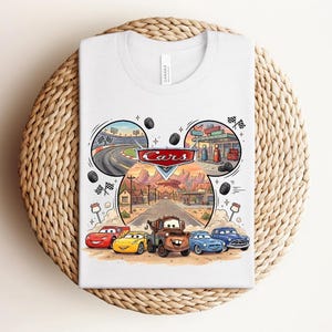 Puede incluir: Una camiseta blanca con un estampado gr&aacute;fico del logotipo de Disney Pixar Cars y personajes como Rayo McQueen, Cruz Ram&iacute;rez y Mate. El dise&ntilde;o incluye escenas circulares de una pista de carreras, una gasolinera y un paisaje de carretera des&eacute;rtica.