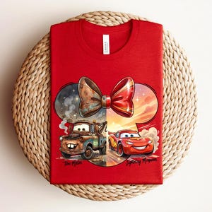 May include: A red t-shirt featuring a graphic of a bow shape containing two cars. On the left, a brown rusty tow truck labelled Tow Mater. On the right, a red race car labelled Lightning McQueen. The design includes smoke and sunset elements.