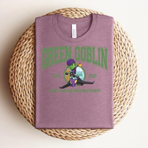 May include: A mauve t-shirt with a crew neck featuring a green graphic of the Green Goblin character riding a glider. The text "GREEN GOBLIN" arches across the top, with "EST 2021" and "ROGUE AND HIS AMAZING FRIENDS" below the image.