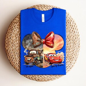May include: A bright blue t-shirt featuring a graphic of Tow Mater and Lightning McQueen from the movie Cars. The design is shaped like a mouse ear silhouette with a large bow on top. The text Tow Mater and Lightning McQueen is printed below.
