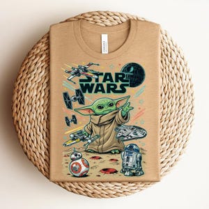 Puede incluir: Una camiseta de color tostado con un estampado gr&aacute;fico que incluye el texto STAR WARS, un personaje alien&iacute;gena verde, una Estrella de la Muerte, un X-wing, cazas TIE, el Halc&oacute;n Milenario, BB-8 y R2-D2, con estrellas y l&iacute;neas de colores.