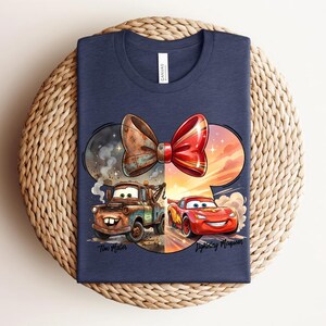 May include: A navy blue t-shirt featuring a graphic of a large bow shape containing two cartoon cars. On the left, a brown tow truck labelled Tow Mater is shown. On the right, a red race car labelled Lightning McQueen is depicted. The design is colourful.