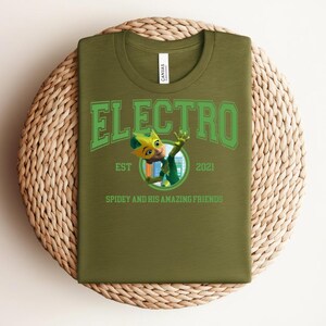 May include: Olive green t-shirt with the word "ELECTRO" in green block letters. Below the word is a cartoon character and the text "EST 2021 SPIDEY AND HIS AMAZING FRIENDS". The shirt is laid on a woven, circular mat.