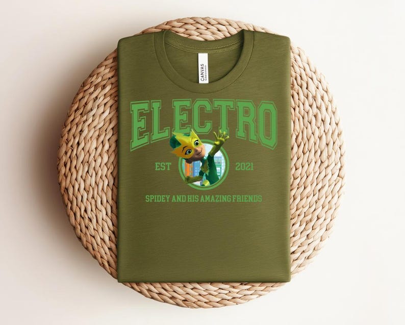 May include: Olive green t-shirt with the word "ELECTRO" in green block letters. Below the word is a cartoon character and the text "EST 2021 SPIDEY AND HIS AMAZING FRIENDS". The shirt is laid on a woven, circular mat.