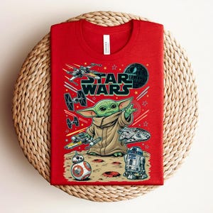 Puede incluir: Una camiseta roja de cuello redondo con un estampado gr&aacute;fico de Star Wars. El dise&ntilde;o incluye a Grogu, BB-8, R2-D2 y varias naves espaciales como el X-wing y el Halc&oacute;n Milenario sobre un fondo espacial con el logotipo de Star Wars en negro.