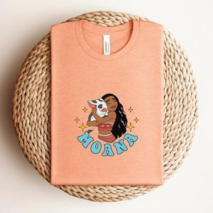 Puede incluir: Una camiseta color melocot&oacute;n con una ilustraci&oacute;n gr&aacute;fica de un personaje de pelo largo y oscuro abrazando a un cerdito. El texto MOANA est&aacute; impreso en azul claro debajo del personaje, rodeado de peque&ntilde;os iconos de estrellas amarillas.