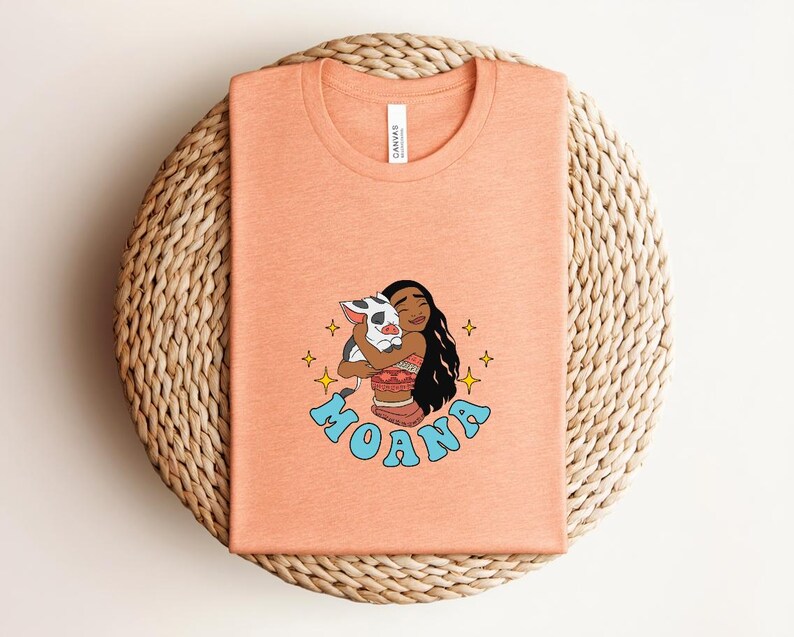 Puede incluir: Una camiseta color melocot&oacute;n con una ilustraci&oacute;n gr&aacute;fica de un personaje de pelo largo y oscuro abrazando a un cerdito. El texto MOANA est&aacute; impreso en azul claro debajo del personaje, rodeado de peque&ntilde;os iconos de estrellas amarillas.