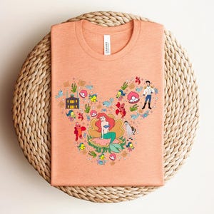 K&ouml;nnte beinhalten: Pfirsichfarbenes T-Shirt mit einer bunten Grafik einer Meerjungfrau und anderer Charaktere. Das Design ist in einer Mickey-Mouse-&auml;hnlichen Form angeordnet. Das Shirt besteht aus einem weichen Material und hat einen Rundhalsausschnitt.