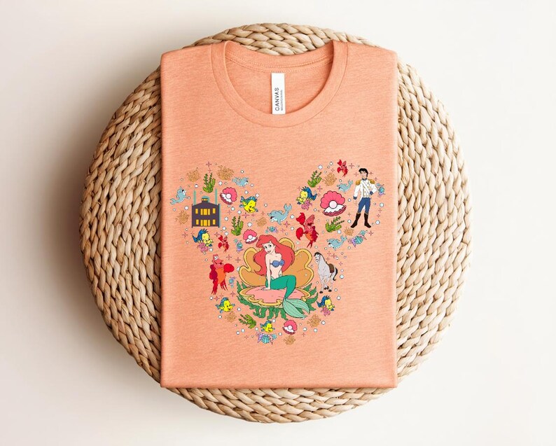 K&ouml;nnte beinhalten: Pfirsichfarbenes T-Shirt mit einer bunten Grafik einer Meerjungfrau und anderer Charaktere. Das Design ist in einer Mickey-Mouse-&auml;hnlichen Form angeordnet. Das Shirt besteht aus einem weichen Material und hat einen Rundhalsausschnitt.
