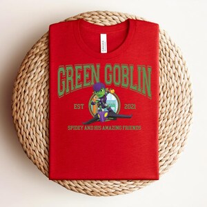 May include: A red t-shirt featuring the text "GREEN GOBLIN" in green arched lettering. Below the text is a cartoon image of the Green Goblin character and the text "SPIDEY AND HIS AMAZING FRIENDS". The shirt is folded on a woven surface.